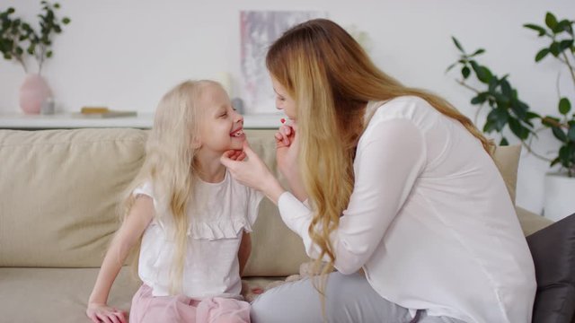 Handheld shot of mother sitting on couch and putting moisturizing lipstick on her adorable little daughter with blond hair
