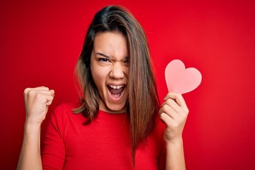 Young beautiful brunette romantic girl holding red paper heart shape over isolated background screaming proud and celebrating victory and success very excited, cheering emotion