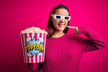Obraz premium Young beautiful girl watching movie using 3d glasses eating box with popcorns smiling cheerful showing and pointing with fingers teeth and mouth. Dental health concept.