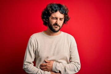 Young handsome man with beard wearing casual sweater standing over red background with hand on stomach because nausea, painful disease feeling unwell. Ache concept.