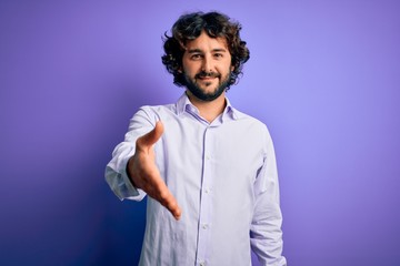 Young handsome business man with beard wearing shirt standing over purple background smiling cheerful offering palm hand giving assistance and acceptance.