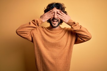 Young handsome man with beard wearing casual sweater standing over yellow background covering eyes with hands smiling cheerful and funny. Blind concept.