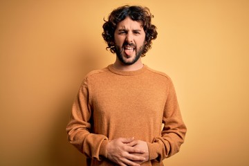 Young handsome man with beard wearing casual sweater standing over yellow background sticking tongue out happy with funny expression. Emotion concept.