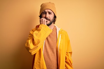 Young handsome man with beard wearing raincoat for rainy day over yellow background looking stressed and nervous with hands on mouth biting nails. Anxiety problem.