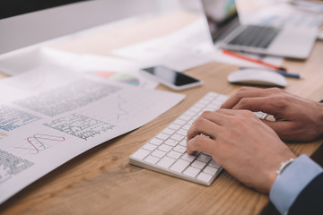 Cropped view of data analyst using computer keyboard near papers with graphs on table