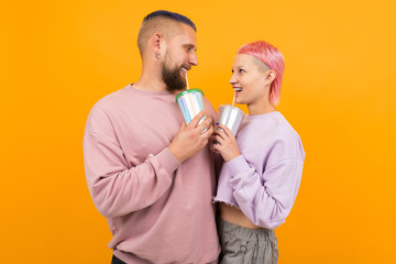 Unusual pretty woman with short pink hair and tattoo drinks coffee and having fun with her boyfriend isolated on orange background
