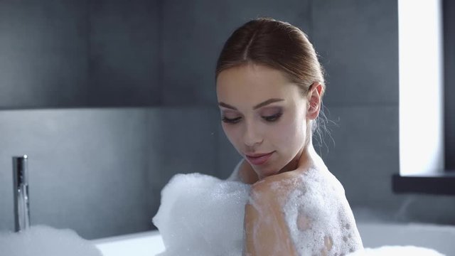 attractive young woman lathering body in bath tub with foam