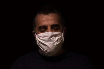 Man wearing white medical face mask as prevention from spreading disease, alone and scared in a dark, eyes closed