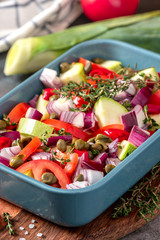 Raw sliced vegetables in a baking dish on the kitchen table. Zucchini, tomatoes, onions, capers and herbs are prepared for baking in the oven. Tasty and healthy vegan and vegetarian food.