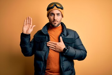 Young handsome skier man with beard wearing snow sportswear and ski goggles Swearing with hand on...