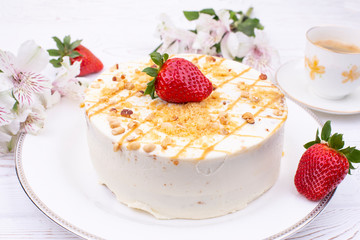 Cake with cream and nuts on a white plate, juicy strawberries and a cup of coffee and flowers on a white shabby table.
