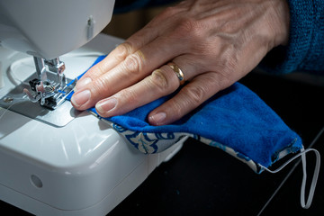 A closeup shows a woman's hand sewing stitches into a homemade covid-19 coronavirus protection mask.