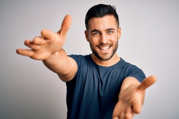 Young handsome man wearing casual t-shirt standing over isolated white background looking at the camera smiling with open arms for hug. Cheerful expression embracing happiness.