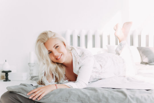 A pretty attractive sexy sensual happy blonde girl with a beautiful smile in underwear and white pajamas is resting, waking up, lying on the bed in a bright room at home, in an apartment