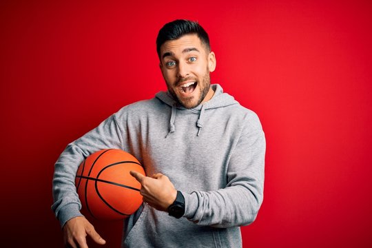 Young sports man holding basketball ball over red isolated background very happy pointing with hand and finger