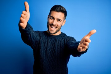 Young handsome man wearing casual sweater standing over isolated blue background looking at the camera smiling with open arms for hug. Cheerful expression embracing happiness.