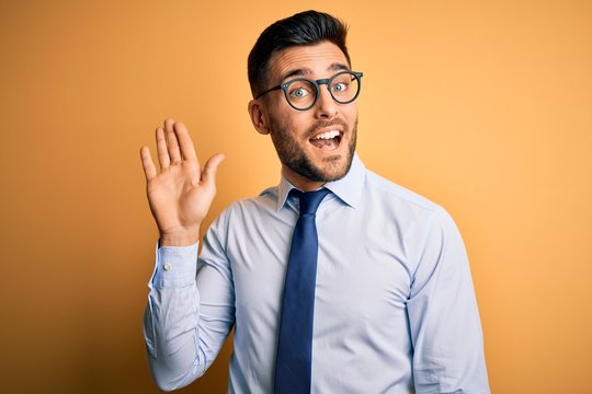 Young handsome businessman wearing tie and glasses standing over yellow background Waiving saying hello happy and smiling, friendly welcome gesture