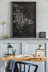 Stylish interior of home office room with black mock up poster map, wooden desk, black chair, clock, books, plants, cacti, office supplies, lamp and personal accessories in modern home decor.