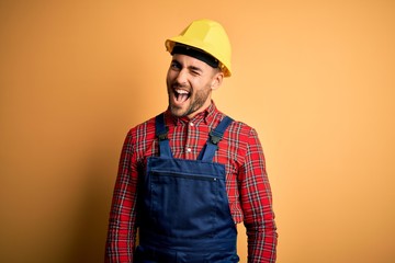Young builder man wearing construction uniform and safety helmet over yellow isolated background...