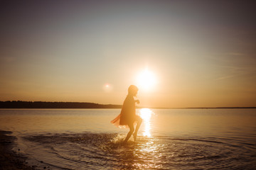 Fototapeta na wymiar Silhouette of a girl running on water against the background of an evening sunset. Water splashes in all directions. Carefree happy baby fun.