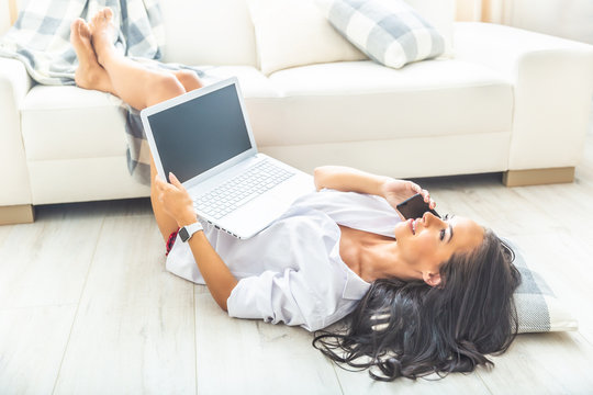 Dark-haired good looking girl lying on her back on the floor with her feet up on a white sofa, a pillow under her head, holding a laptop and having a phone call