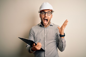 Young architect inspector man wearing builder safety helmet and using clipboard very happy and...