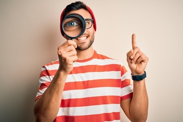 Young detective man looking through magnifying glass over isolated background surprised with an idea or question pointing finger with happy face, number one
