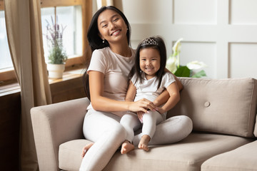 Obraz premium Happy motherhood, loving family, parental protection, life value concept image. Asian young mother and cutie little daughter in casual home clothes seated on couch embracing smiling looking at camera