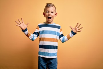 Young little caucasian kid with blue eyes wearing colorful striped shirt over yellow background celebrating crazy and amazed for success with arms raised and open eyes screaming excited. Winner 