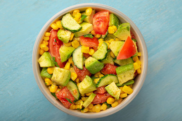 Top view bowl of avocado and tomato salad with cucumber and corn on a blue background.