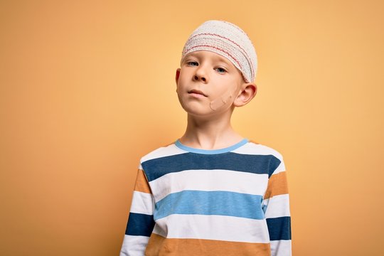 Young little caucasian kid injured wearing medical bandage on head over yellow background Relaxed with serious expression on face. Simple and natural looking at the camera.