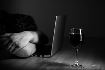Home office and alcoholism problem - depressed man sitting in front of computer, he hide his face. Wine glass on the table