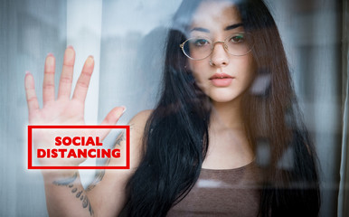 Social distancing sign on teen girl portrait through window at home due to coronavirus