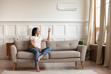 Young asian woman switching on air conditioner while resting seated on couch in modern interior...