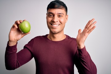 Young hispanic man eating fresh green apple as healthy nutrition over isolated background very happy and excited, winner expression celebrating victory screaming with big smile and raised hands