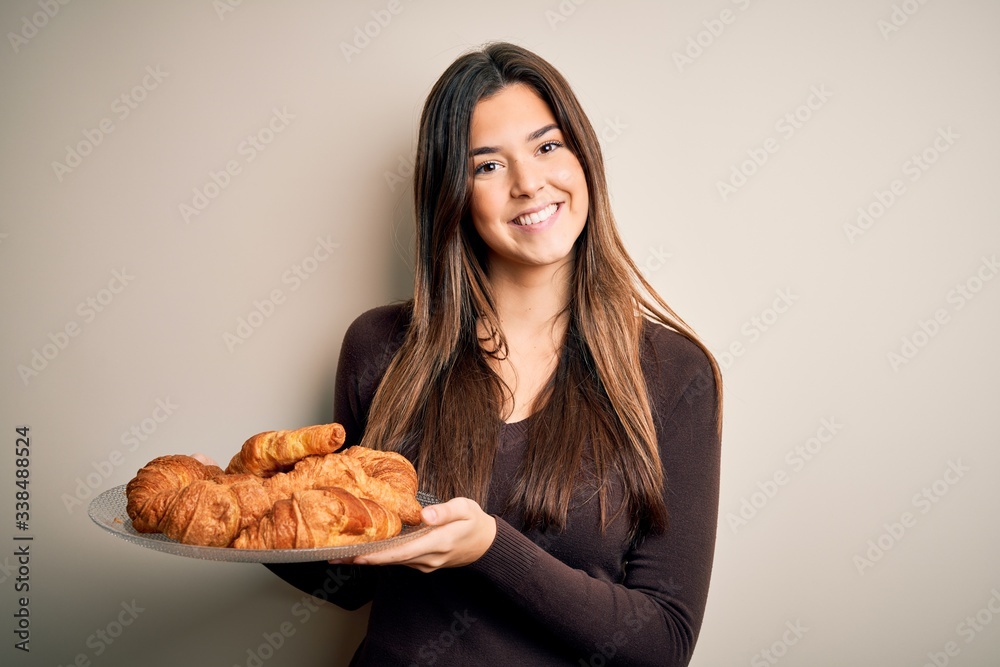 Poster Young beautiful girl holding plate with sweet croissants for breakfast over white background with a happy face standing and smiling with a confident smile showing teeth