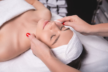 Young woman is enjoying facial procedure at beauty salon. Girl is lying in spa and getting face massage with pleasure