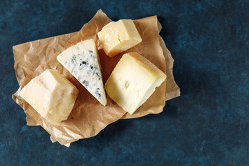 Different sorts of cheese on a parchment paper on dark blue textured background. Top view