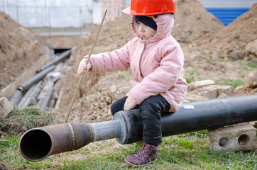 a child in a protective helmet sits on a pipe on a heating main and  fixing welding and grinding industrial construction oil and gas or water plumbing pipeline outside on site