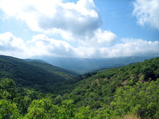 Valley of hills covered with low green forest. In the distance, the silhouettes of high mountains turn blue. Clear blue sky with small clouds.