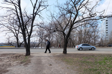 KYIV, UKRAINE - MARCH 18, 2015: Old man walking on the ground among two geometrical trees at empty street. Road with the light blue car and building on the background