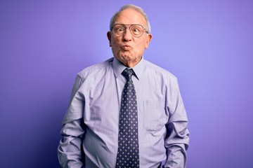 Grey haired senior business man wearing glasses standing over purple isolated background making fish face with lips, crazy and comical gesture. Funny expression.