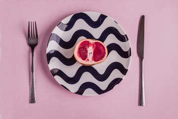 grapefruit on a striped plate with a knife and fork on a pink background, morning breakfast