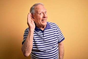 Grey haired senior man wearing casual navy striped t-shirt standing over yellow background smiling with hand over ear listening an hearing to rumor or gossip. Deafness concept.