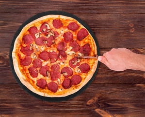 Salami pizza with mushrooms, on slate bottom, on wooden table hand cutting pizza, top view