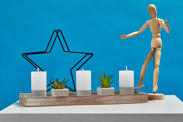 Fototapeta na wymiar White table, figurine of human, green plants in pots, three big candles and one small in candlestick in form of iron star. Blue background. Close up