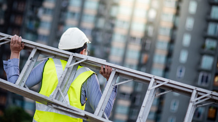 Asian maintenance worker man with safety helmet and green vest carrying aluminium step ladder at construction site. Civil engineering, Architecture builder and building service concepts © zephyr_p
