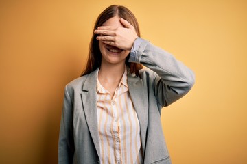 Young beautiful redhead woman wearing jacket and glasses over isolated yellow background smiling and laughing with hand on face covering eyes for surprise. Blind concept.