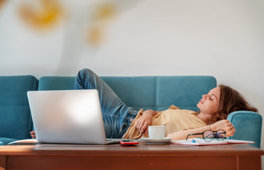 Charming beautiful young girl in glasses with curly hair fell asleep on a blue sofa at home in front of a laptop, notepad and a cup of coffee, remote work and education. Fatigue, Deadline