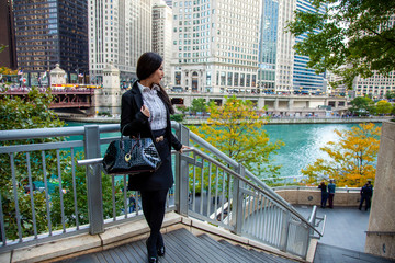 Beautiful Chinese Asian woman in fashionable business attire is enjoying a nice day walking around downtown Chicago in the afternoon.  She carries a trendy handbag and black blazer with red lipstick - obrazy, fototapety, plakaty